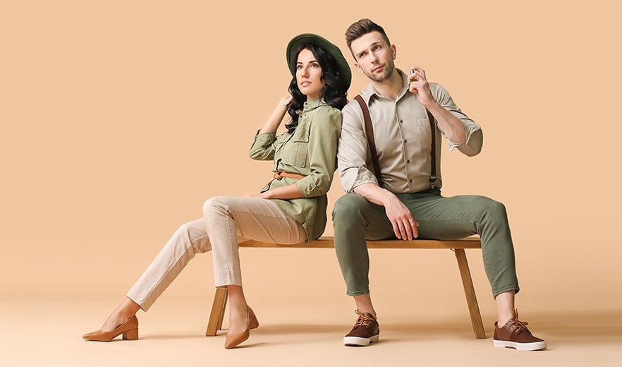 Ein Mann und eine Frau in modischen Klamotten posieren auf einer Holzbank vor einem braunen Hintergrund